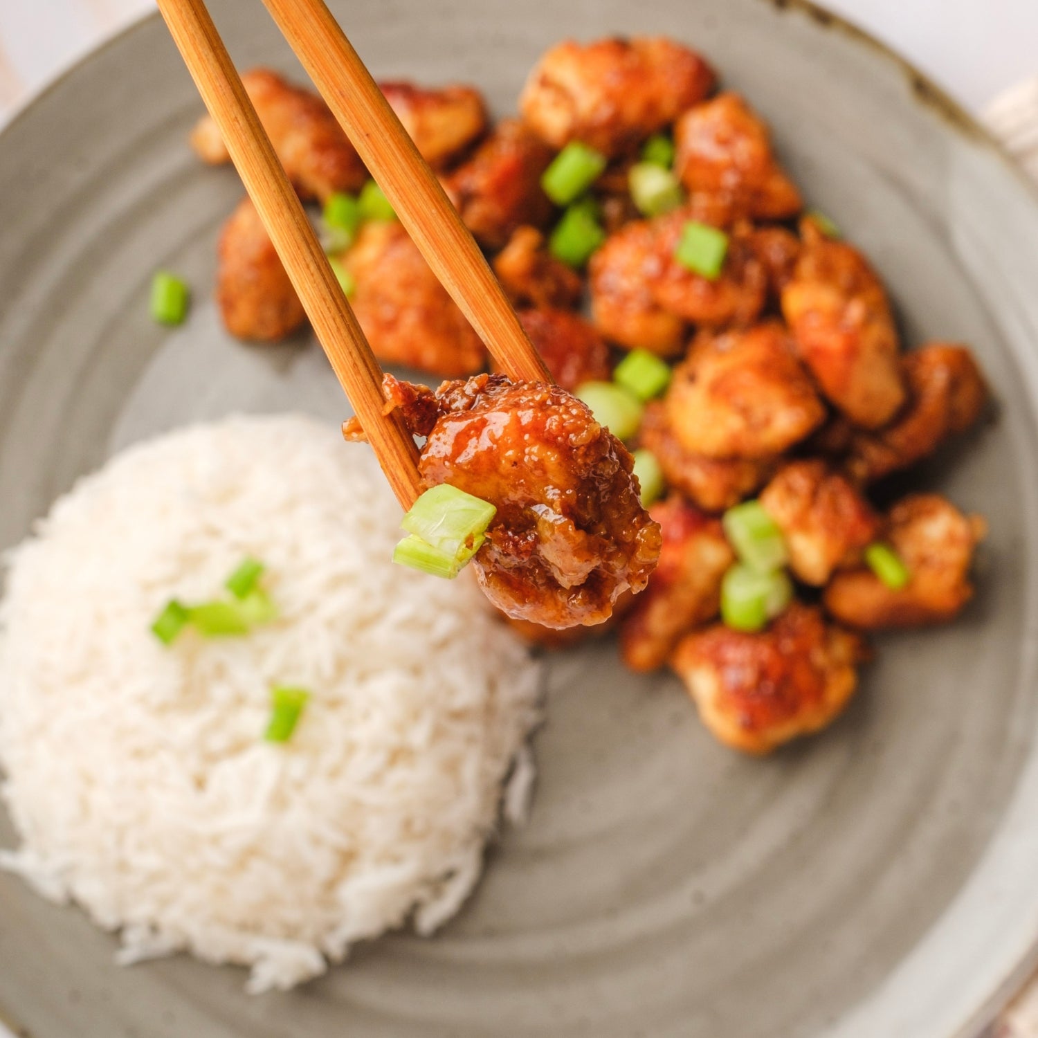 Honey chicken being held by chopsticks with chicken and rice in the background.