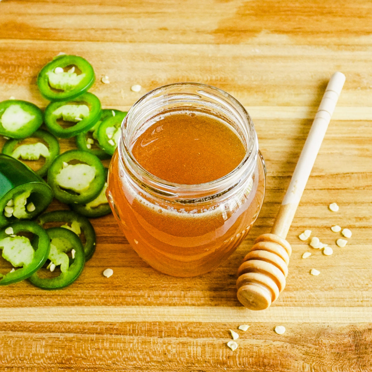 Hot honey in a jar next to a honey stick and chopped jalapenos, all on top of a cutting board.