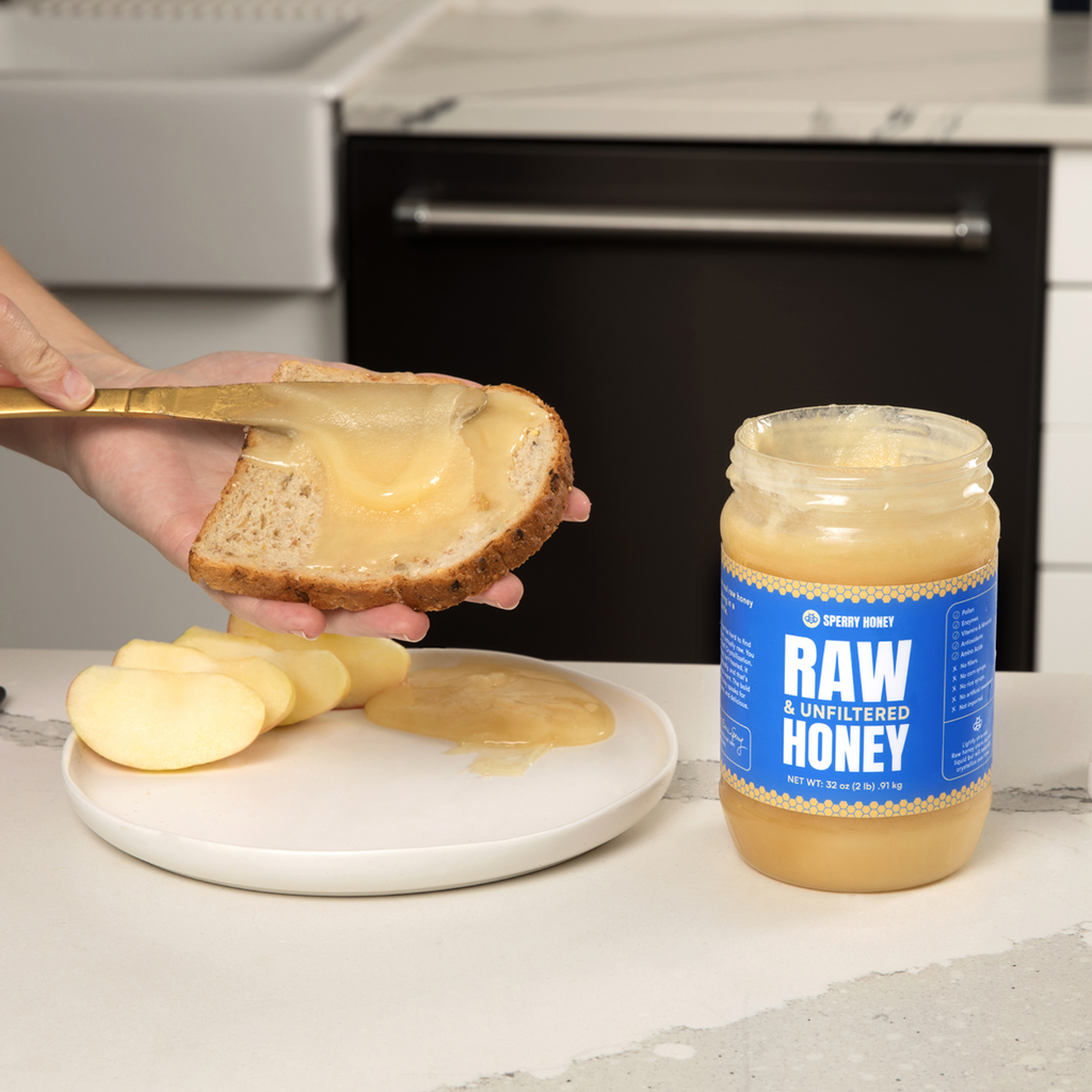 Person spreading honey on toast with a jar of honey labeled 'Raw & Unfiltered Honey' in the foreground.