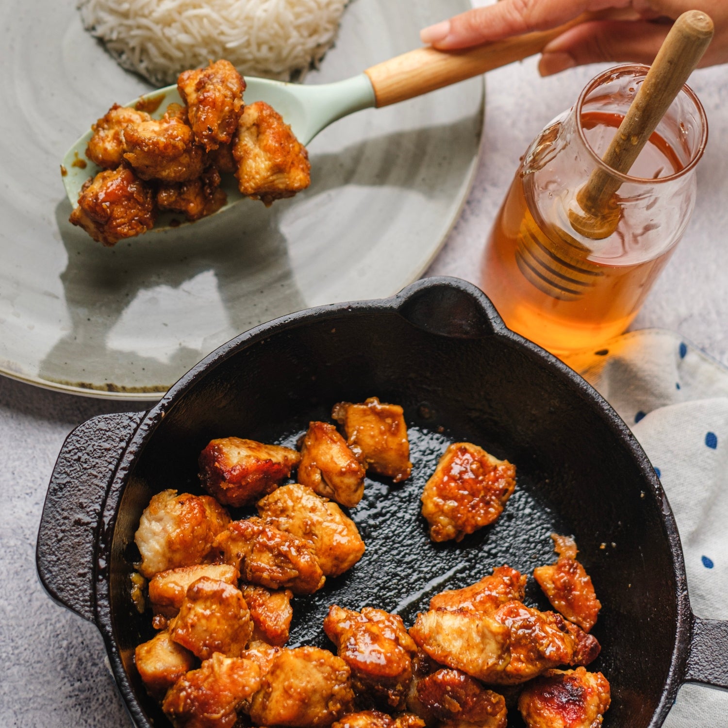 Someone scooping honey chicken onto a plate next to a jar of honey and a skillet of chicken.