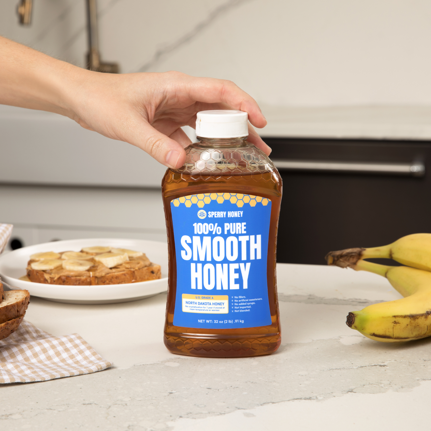 Bottle of Sperry Honey with a hand holding it, surrounded by bananas and toast on a kitchen counter.